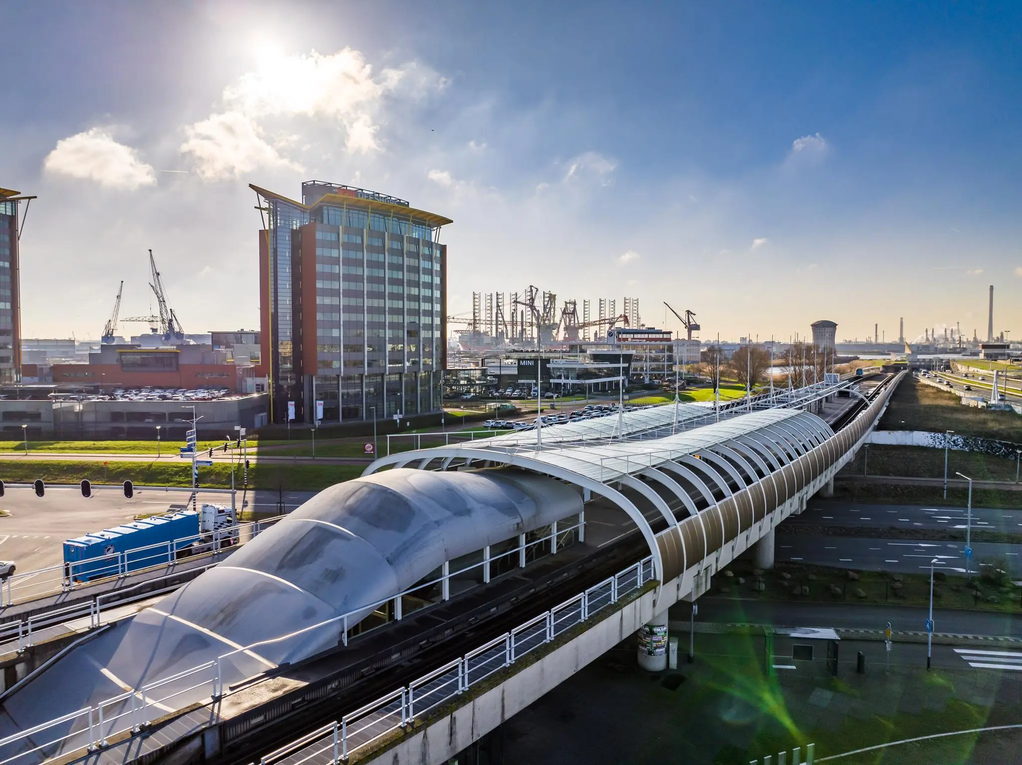 Luchtfoto van de Karel Doormanweg in Rotterdam met een modern metrostation, kantoorgebouwen en de havenkranen op de achtergrond.